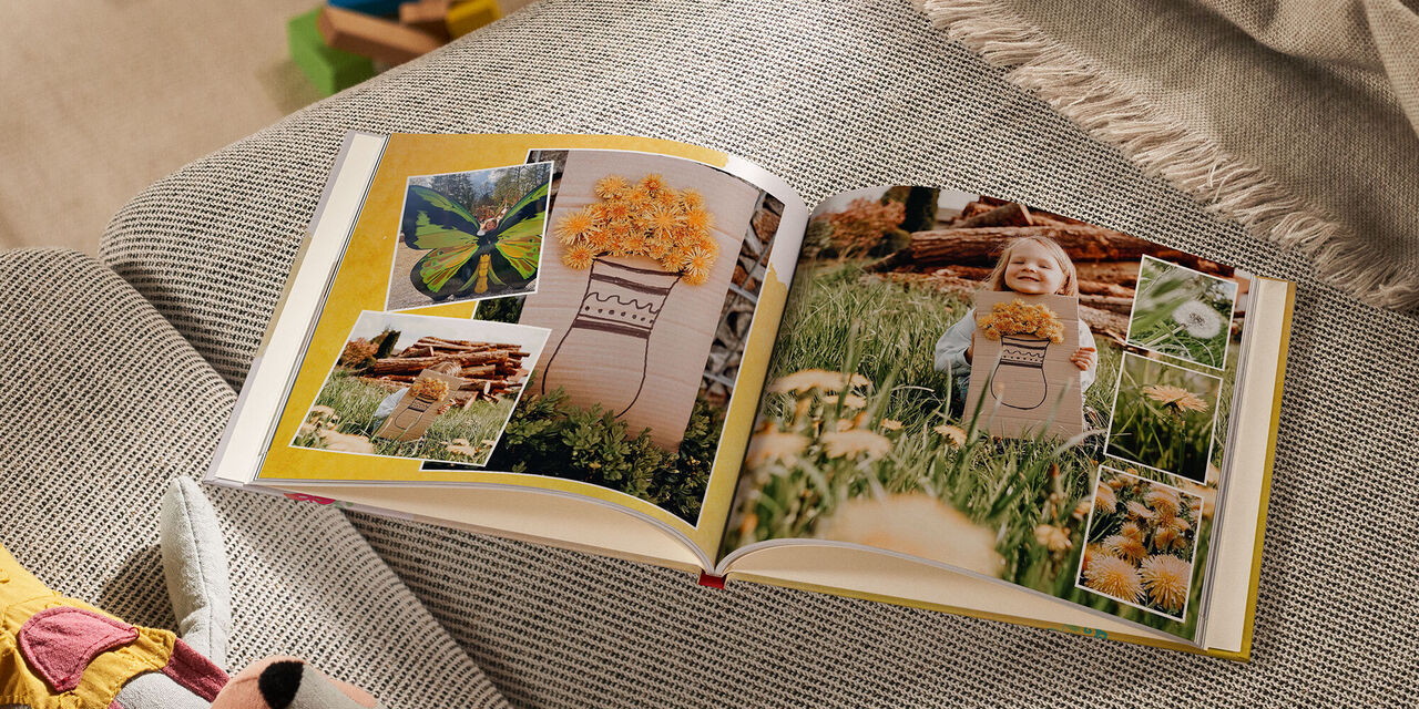 Un FOTOLIBRO CEWE aperto è appoggiato su un divano accanto a un peluche e a una coperta. Sul lato sinistro ci sono tre foto, due delle quali mostrano il lavoro artigianale di una bambina: dei fiori di dente di leone sono stati incollati su un pezzo di cartone, sul quale è stato dipinto anche un vaso di fiori. La terza immagine mostra grandi ali di farfalla con una bambina in piedi dietro di esse. La pagina di destra è completamente occupata da una foto della bambina con il suo lavoro artigianale. Tre foto più piccole sulla stessa pagina mostrano i dettagli del dente di leone come fiore e come soffione.