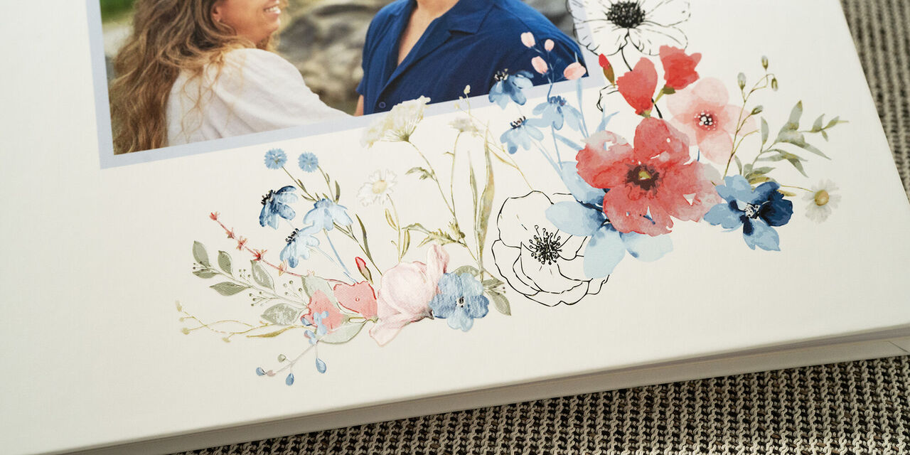 Puoi vedere un primo piano della copertina del libro degli ospiti. Mostra una foto della coppia di sposi. Sotto c’è un design floreale da matrimonio con finitura laccata su sfondo bianco.